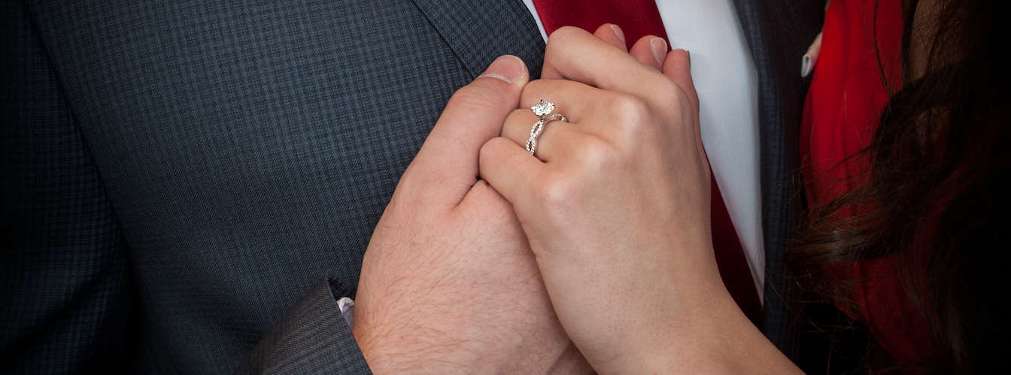 Holding hands. Woman wears a cushion cut diamond ring.